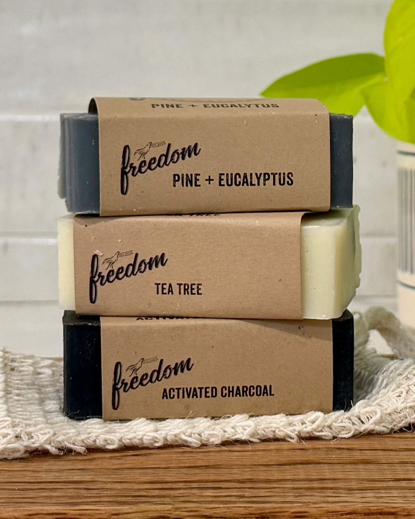 Three bars of soap with Freedom Soap wraps labeled 'pine + eucalyptus', 'tea tree', and 'activated charcoal' on a wooden surface.