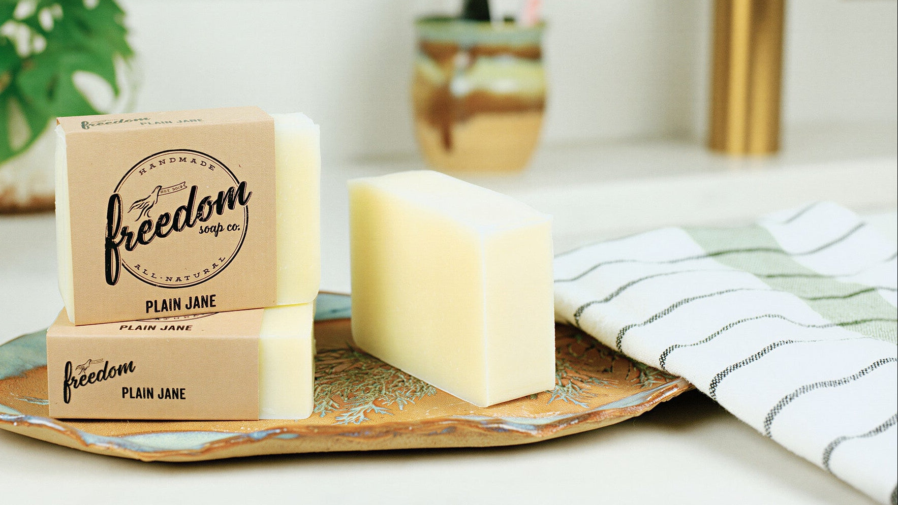 Two bars of Plain Jane soap by Freedom Soap Co. displayed on a decorative tray on a bathroom counter, with a striped towel, toothbrush holder, and faucet in the background.
